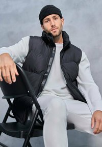 Gilet rembourré noir avec un col haut, porté sur une chemise blanche à manches longues texturée. Modèle assis sur une chaise pliante noire devant un fond gris.