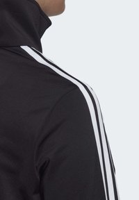 Veste noire à col montant avec des accents rayés blancs le long des épaules, dotée d'une texture lisse et extensible et d'un design minimaliste.