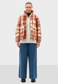 Oversized red and cream checkered fleece jacket with two front pockets, paired with a white shirt and blue wide-leg pants. Black ankle boots.