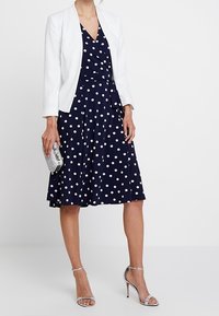 Granatowa sukienka w groszki z kopertowym fasonem, zestawiona z białą marynarką. Stylizację uzupełniają srebrne sandały na wysokim obcasie i metaliczna kopertówka.