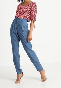 Blusa de estampado rojo con mangas cortas, combinada con pantalones de tiro alto azules y tacones descalcificados. Texturas suaves y formas definidas.