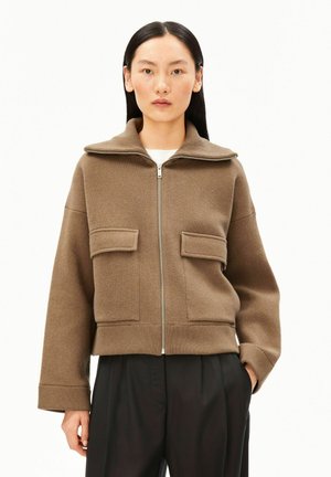 Femme portant une veste marron zippée avec de grandes poches avant et un large col, associée à un pantalon noir, debout sur un fond blanc.