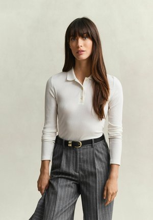Junge Frau mit langen braunen Haaren, die ein weißes, geripptes Langarm-Polo und graue Nadelstreifenhose mit schwarzem Gürtel trägt.