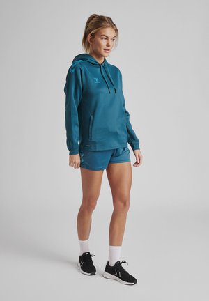 Türkise Sportkapuzenjacke mit Kängurutasche und schwarzen Kordeln, kombiniert mit passenden Shorts. Das Model trägt schwarze Sneakers und weiße Knöchelsocken.