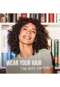 Lachende vrouw met krullend haar staat voor een boekenplank, draagt een lichtgrijze trui, met de tekst "Draag je haar zoals jij wilt."