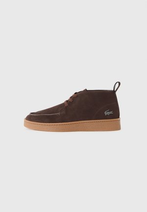 Baskets montantes en suède marron avec fermeture à lacets, semelle en gomme, languette au talon et petit logo crocodile sur le côté sur fond blanc.