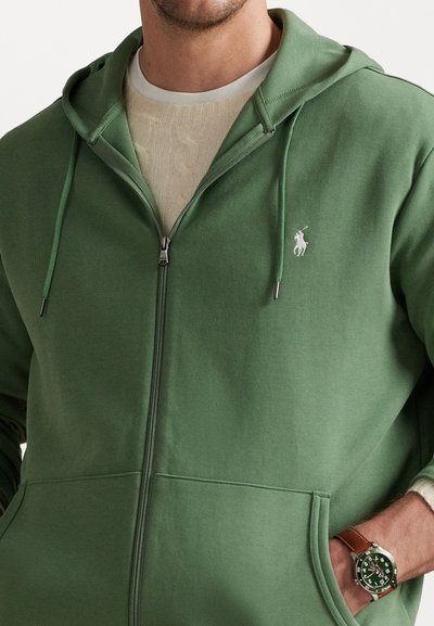 Grön zip-up hoodie med snörning på huven, med en vit logotyp på bröstet och en känguruficka. Under finns en krämfärgad, texturerad tröja synlig.