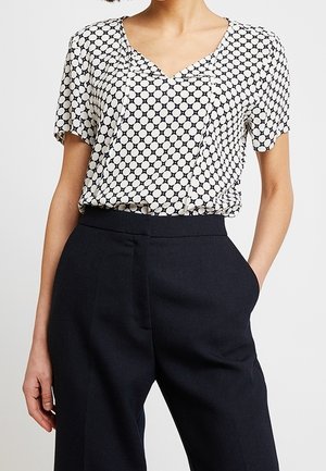 Blusa corta de manga corta en blanco y negro con un patrón de lunares, con escote en V y tejido ligero, combinada con pantalones negros ajustados.