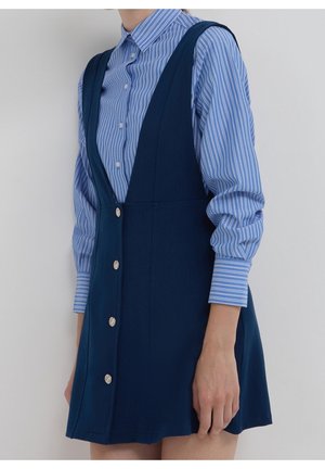Vrouw draagt een blauw gestreept overhemd met knopen onder een mouwloze marineblauwe jurk met grote zilveren knopen aan de voorkant.