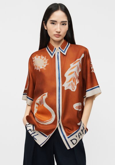 Νεαρή γυναίκα με μακριά μαύρα μαλλιά που φοράει μια φαρδιά, κοντομάνικη πορτοκαλί πουκάμισο με μπλε και λευκή λεπτομέρεια και μαύρο παντελόνι.