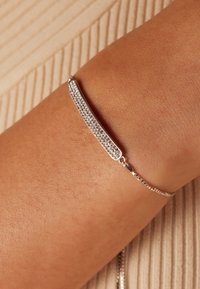 Silberarmband mit einem gebogenen Balken, der eine Reihe kleiner klarer Steine aufweist, verbunden mit einer zarten Kette, die auf einer strukturierten Hautoberfläche ruht.