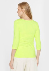 Neon-gelbes, enganliegendes Langarmoberteil mit rundem Ausschnitt. Der Stoff ist glatt und dehnbar und hat Ärmel in Dreiviertellänge.