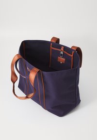 Borsa a tote in tela di un viola scuro con dettagli e manici in pelle marrone. L'interno presenta cuciture a contrasto e una piccola tasca con chiusura.