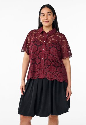Bordowa koszula koronkowa z kwiatowym wzorem, krótki rękaw, kołnierzyk i falbaniasty dół, w zestawie z czarną spódnicą plisowaną.
