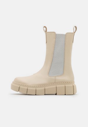 Botte Chelsea beige chunky avec semelle épaisse rainurée, panneau latéral élastique et languettes de tirage sur fond blanc, vue de côté.