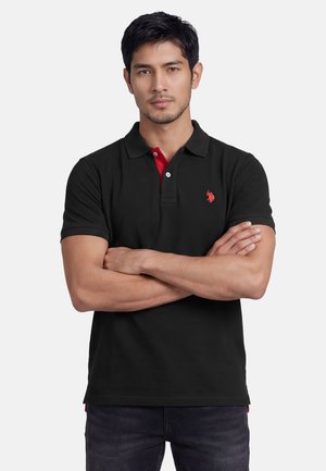 Man with folded arms wearing a black polo shirt with red logo and red accents, standing against a plain light background.