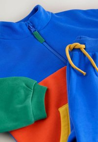 Zils rāvējslēdzēja tops ar zaļiem, oranžiem un dzelteniem akcentiem; ribota aproces un jostasvieta; mīksts, gluds audums ar ērtu piegriezumu.