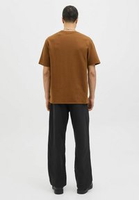 Camisa marrón de manga corta con un acabado texturizado, combinada con pantalones negros de corte holgado, vista desde la parte trasera contra un fondo liso.