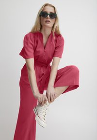 Tuta rossa con finitura testurizzata, maniche corte, cintura con nodo e dettagli in bottone. Abbinata a sneakers bianche alte e occhiali da sole verdi.