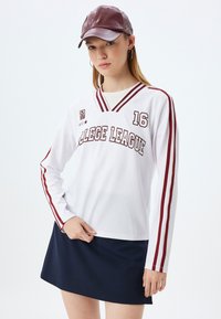 Weißes Langarmtrikot mit roten Streifen, mit dem Text "COLLEGE LEAGUE" und der deutlich sichtbaren Nummer "16". Kombiniert mit einem navyrock und einer braunen Kappe.