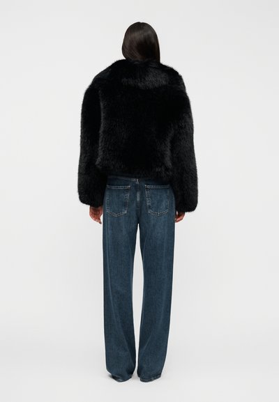 Μαύρο μπουφάν από συνθετική γούνα με κοντό σχεδιασμό, παχύ υφή και φαρδιά μανίκια, συνδυασμένο με άνετο τζιν παντελόνι σε μπλε απόχρωση.