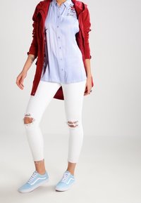 Röd lätt jacka över en ljusblå skjorta med knappar, vita slitna skinny jeans och ljusblå sneakers med vita skosnören.