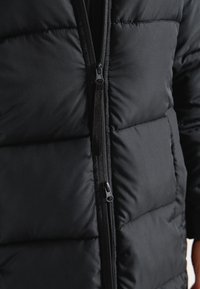 Svart vadderad jacka med en framdragkedja, kviltat mönster och slät tyg. Framträdande detaljer inkluderar en kontrasterande dragkedja och sömmar.