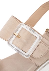 Primer plano de la tira de una sandalia de ante beige con una hebilla cuadrada transparente y un pasador dorado sobre un fondo blanco.