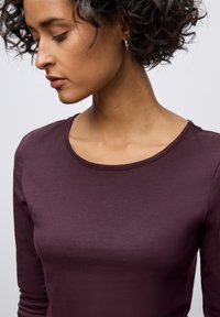 Long-sleeve top in deep burgundy fabric, featuring a rounded neckline and fitted shape. Smooth texture with subtle sheen.