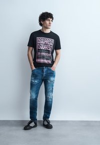 Muž s kučeravými vlasmi v čiernom tričku s potlačou DSQUARED2, opotrebovaných modrých džínsach a čiernych teniskách, stojaci pred jednoduchou svetlou stenou.