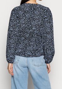 Svart blus med blå och vita spridda mönster, med långa puffärmar och elastisk smock vid midjan. Bärs med ljusblå jeans.