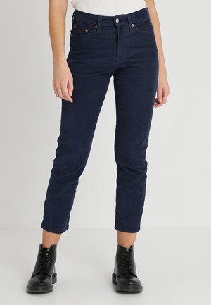 Kvinde iført mørkeblå slim-fit bukser og sorte snørestøvler ved anklen, stående mod en ensfarvet lys baggrund.
