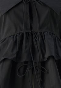 Svart topp med volangkant, bred krage og justerbare bånd. Laget av et glatt stoff med en teksturert overflate.