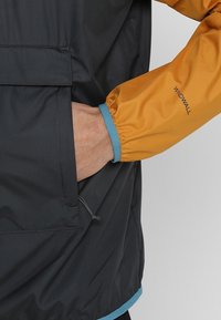 Main dans la poche d'une veste noire avec une manche jaune moutarde portant l'étiquette Windwall et un poignet élastique bleu.