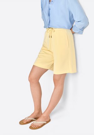 Pantaloni corto giallo chiaro con vita regolabile, dotati di tasche laterali e vestibilità comoda. Abbinati a infradito marroni e una camicia azzurra con bottoni.