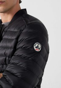 Chaqueta negra acolchada con diseño de pespuntes y cuello ajustado. Cuenta con un parche redondo con el logotipo en el hombro izquierdo con el texto "JOTT".