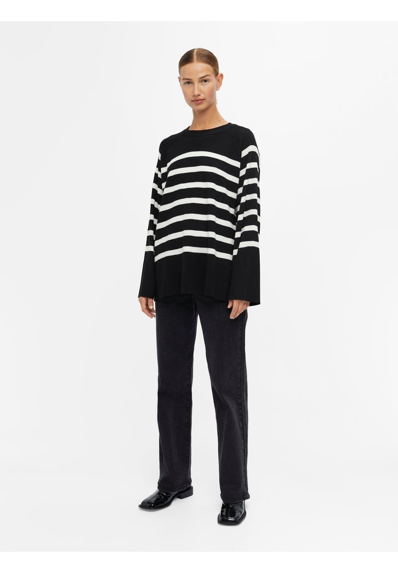 Pull oversized à rayures noires et blanches avec des manches longues, associé à un jean droit noir et des chaussures noires brillantes.