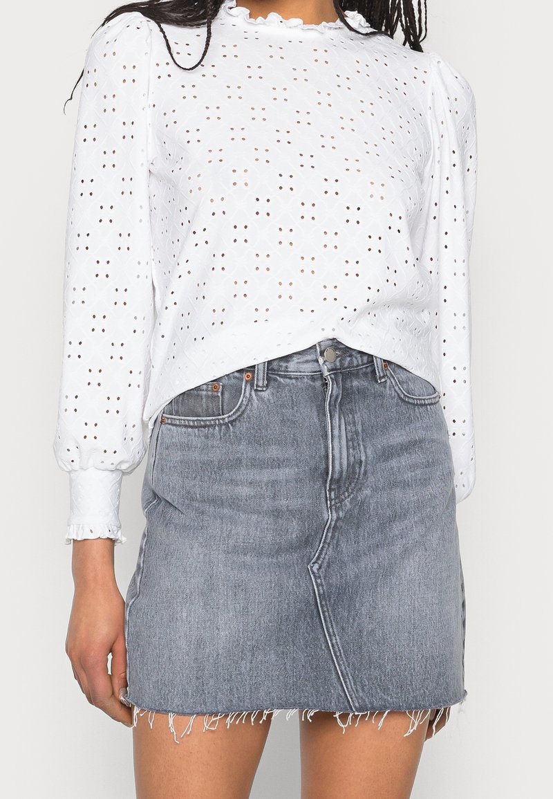 Femme portant un chemisier blanc à manches longues en broderie anglaise avec col et poignets à volants, associé à une mini-jupe en jean gris taille haute avec ourlet effiloché.