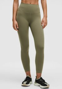 Egy nő magas derékú olívazöld leggingset és fekete sportcipőt visel, egy sima világos háttér előtt áll.