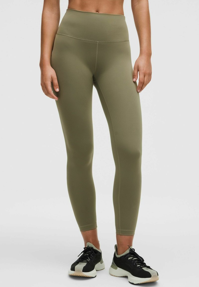Kvinna klädd i högmidjade, olivgröna leggings och svarta träningsskor, stående mot en enkel ljus bakgrund.