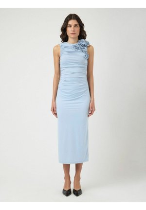 Naine seisab näoga edasi, seljas varrukateta helesinine voltidega midi kleit, millel on kolm kangast lille kaunistust parema õla lähedal, mustad kontsakingad.