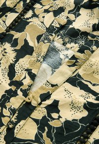 Tissu à motif floral dans des tons de noir, beige et crème, avec un devant à boutons et des accents texturés le long de la patte de boutonnage.