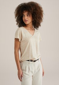 Beige, rövid ujjú felső V-nyakkal, texturált anyagból. Világos színű nadrággal és fekete övvel, arany csattal kombinálva.