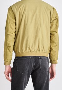 Bomber bunda olivově zelená s žebrovaným lemem a rukávy, s hladkou texturou a zipovou kapsou na levém rukávu.