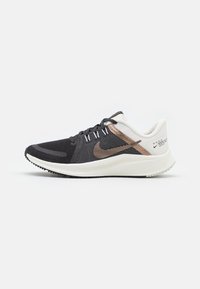 Zapatilla de running en blanco y negro con parte superior de malla, acentos de bronce, diseño de cordones y suela de goma texturizada. Presenta el logo de Nike en el costado.