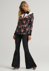 Blusa floral de manga comprida em preto e rosa com colarinho branco, combinada com jeans escuros flare e sapatos castanhos de salto.