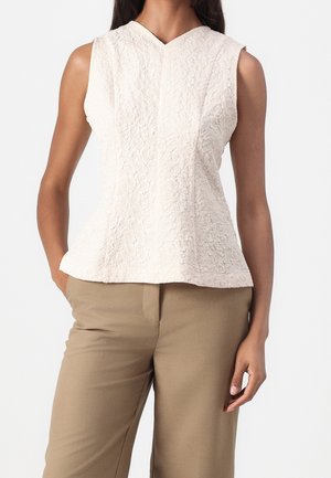 Woman wearing sleeveless white textured top with v-neck paired with high-waisted beige pants, standing with one hand in pocket.