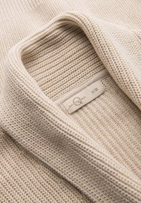 Strickpullover in hellbeige, mit einem strukturierten Muster und einem umgeschlagenen Kragen. Etikett sichtbar, das Größen- und Markendetails anzeigt.