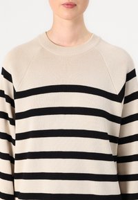 Beige og svart stripete genser med rund hals og ribbestruktur. Har raglanermer og horisontale striper i ulik bredde.