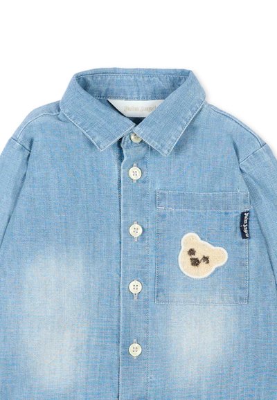 Chemise en denim bleu clair à boutons avec une petite poche ornée d'un patch avec un visage d'ours crème et une étiquette de marque noire sur le côté droit.